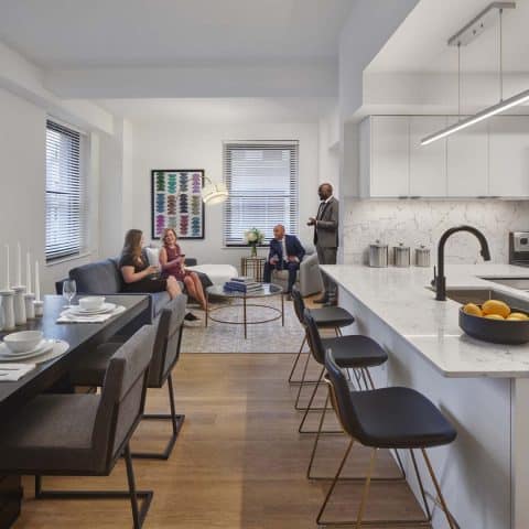 The kitchen and dining room at 101 dupont place. The dining room table is set out with white dishes and placemats and candles. The kitchen features a breakfast bar with marble counters and white cabinets, and stainless steel appliances. in the background guests are mingling and smiling in the living room